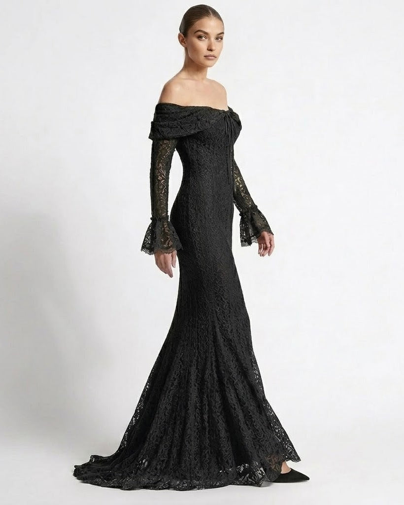 A woman modeling a black off-the-shoulder lace evening gown with long flared sleeves and a fitted mermaid silhouette, photographed against a plain light gray studio background.