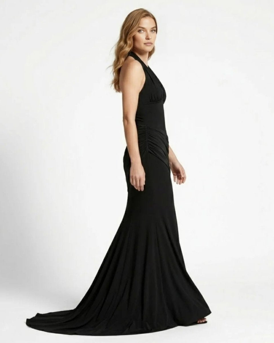 A woman modeling a floor-length black halter gown with a fitted silhouette and draped skirt, photographed against a plain light gray studio background.