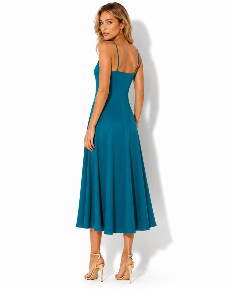 A woman modeling a teal blue midi dress from the back, featuring thin spaghetti straps, a fitted bodice, and a flared skirt, paired with beige high-heeled sandals against a white studio background.