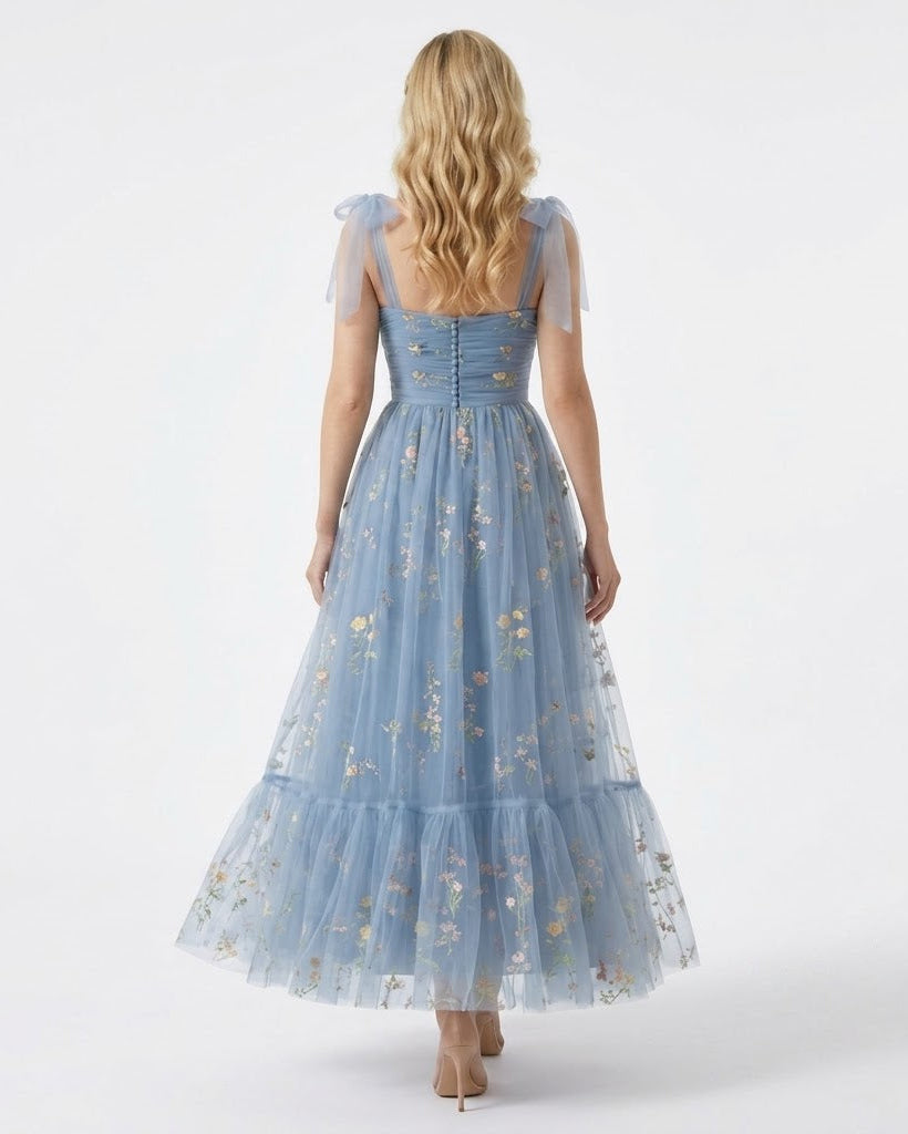 A woman viewed from behind wearing a light blue tulle maxi dress with sheer flutter sleeves, a corset-style back, and delicate floral embroidery, set against a plain white studio background.