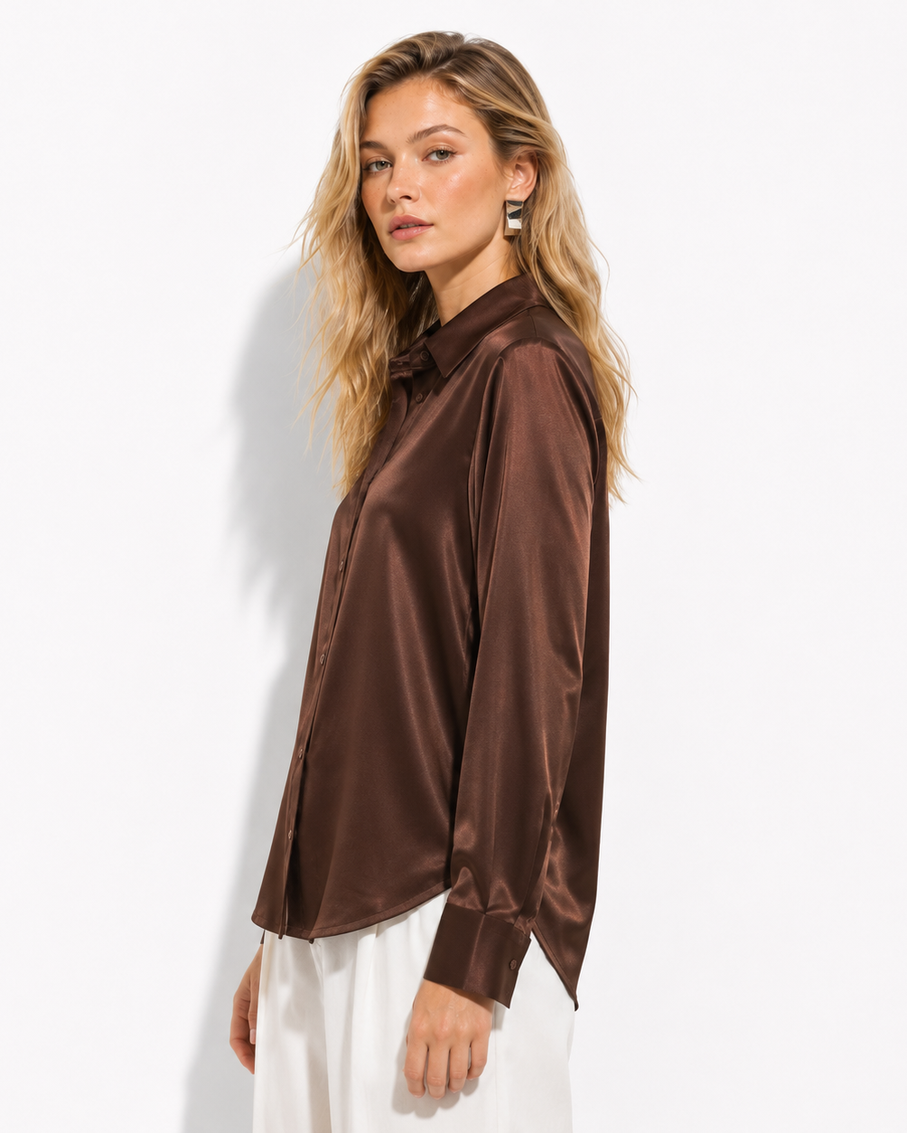 A woman modeling a long-sleeve satin-like brown button-up shirt with a relaxed fit, paired with white pants, against a clean white studio background.