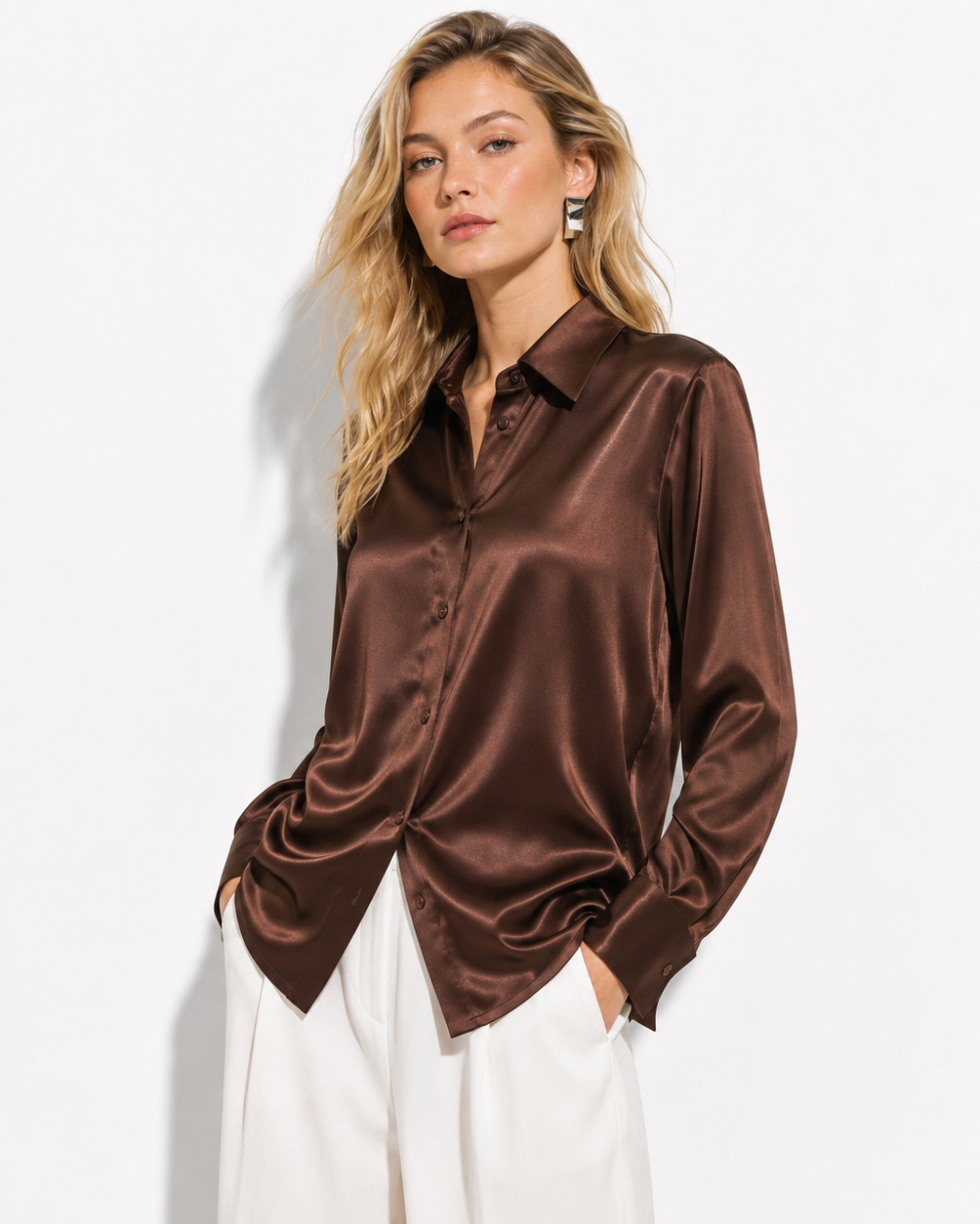 A woman wearing a brown satin button-up shirt and white pants, posing against a clean white studio background.