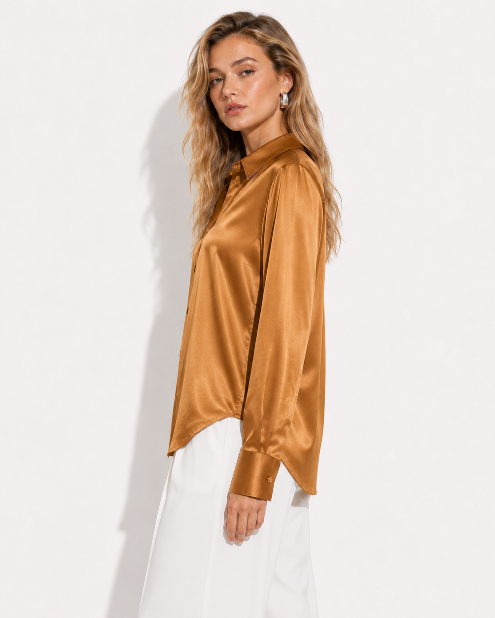A woman modeling a long-sleeve satin blouse in a warm camel brown color, paired with white trousers, against a plain white studio background.