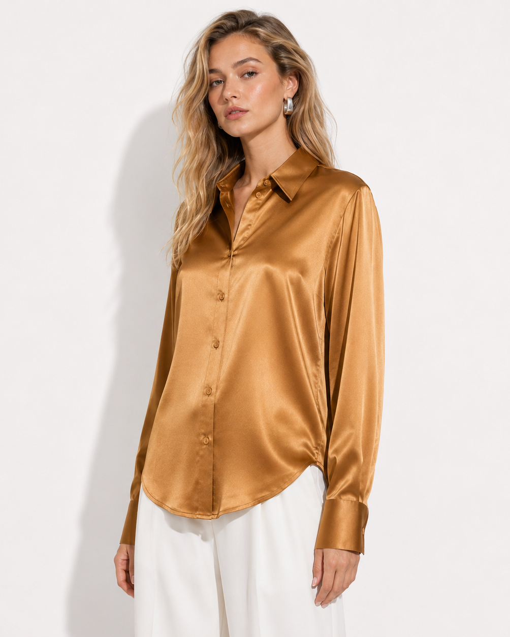 A woman modeling a long-sleeve satin button-up blouse in a warm metallic gold color, paired with white trousers, against a plain white studio background.