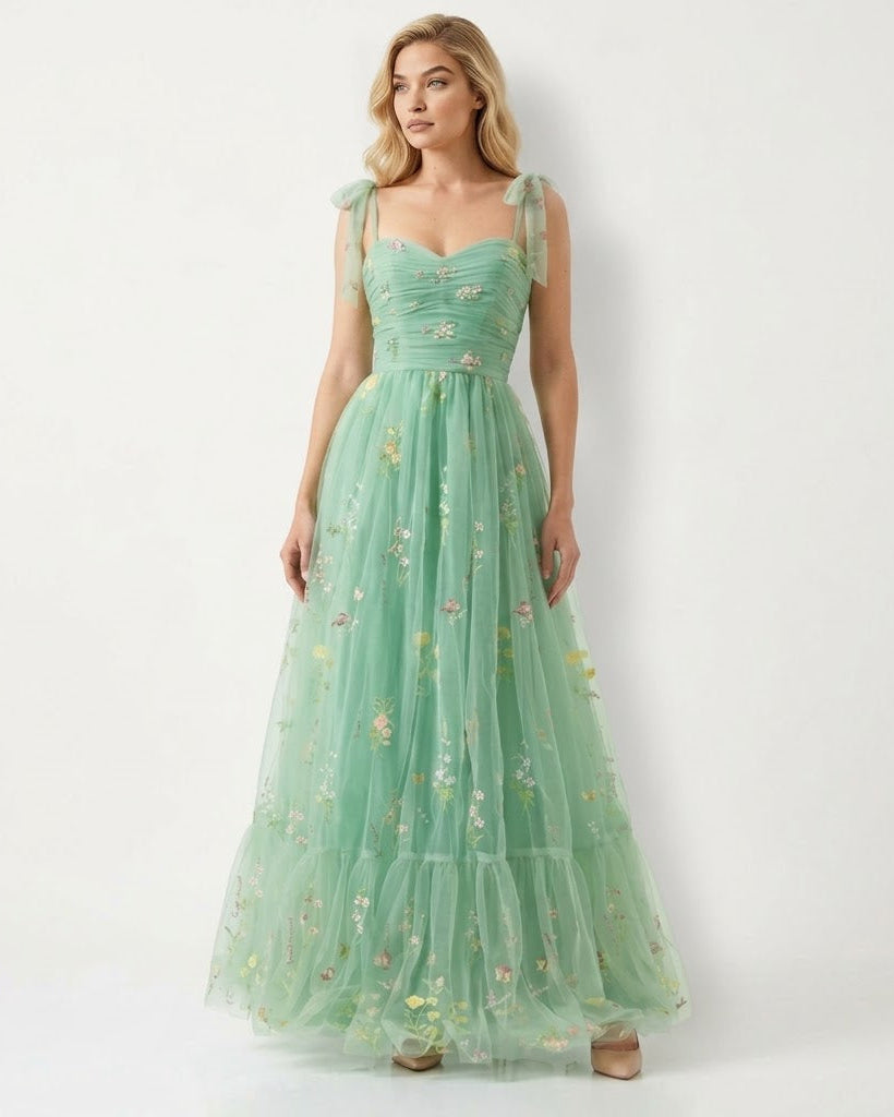 A woman modeling a long mint green tulle dress with floral embroidery and sheer shoulder straps against a plain white studio background.