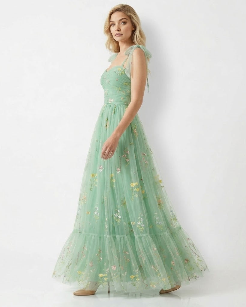 A woman modeling a long, flowing mint green tulle maxi dress with delicate floral embroidery and sheer shoulder straps, set against a plain white studio background.