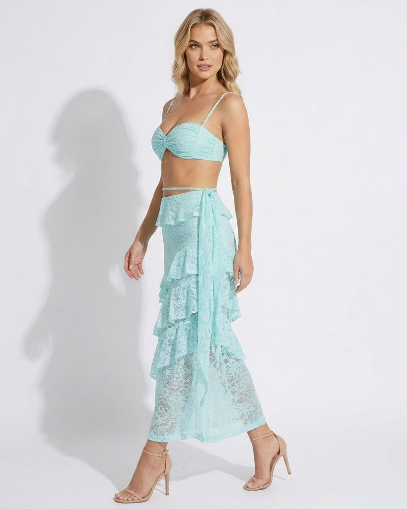 A woman modeling a light aqua two-piece set consisting of a lace bandeau crop top and a matching tiered ruffle maxi skirt, paired with nude high-heeled sandals against a white studio background.