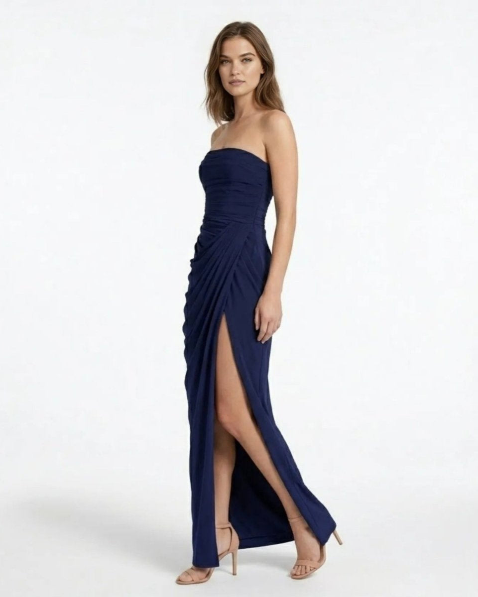 A woman modeling a strapless navy blue maxi dress with a ruched bodice, draped skirt, and high front slit, paired with nude high-heeled sandals against a plain white studio background.