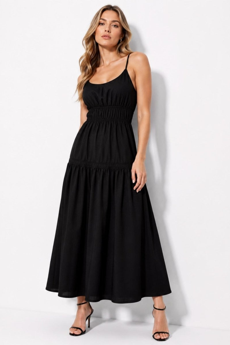 A woman modeling a black midi-length spaghetti-strap maxi dress with a smocked waist and tiered skirt, paired with black strappy heels, against a plain white studio background.