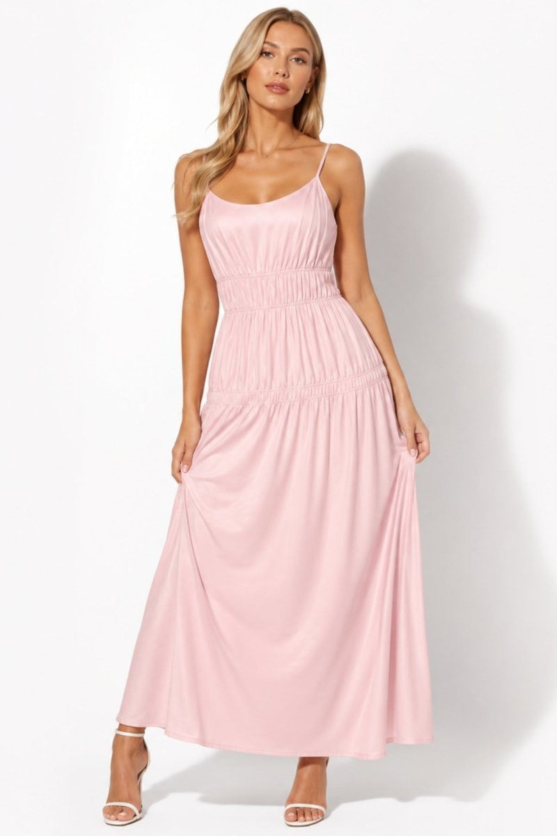 A woman modeling a light pink sleeveless maxi dress with thin spaghetti straps, a smocked bodice, and a gathered skirt, paired with white heeled sandals against a white studio background.