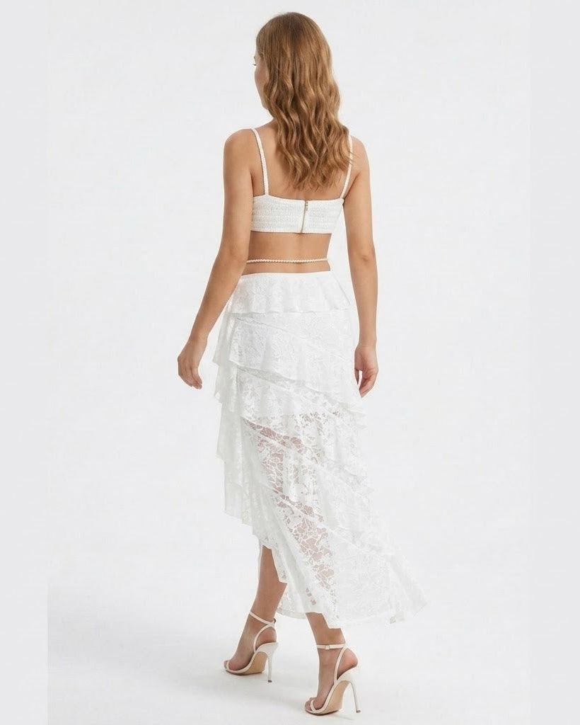 A woman shown from the back wearing a white two-piece outfit consisting of a spaghetti-strap crop top and a tiered lace midi skirt, paired with beige high-heeled sandals against a plain white studio background.
