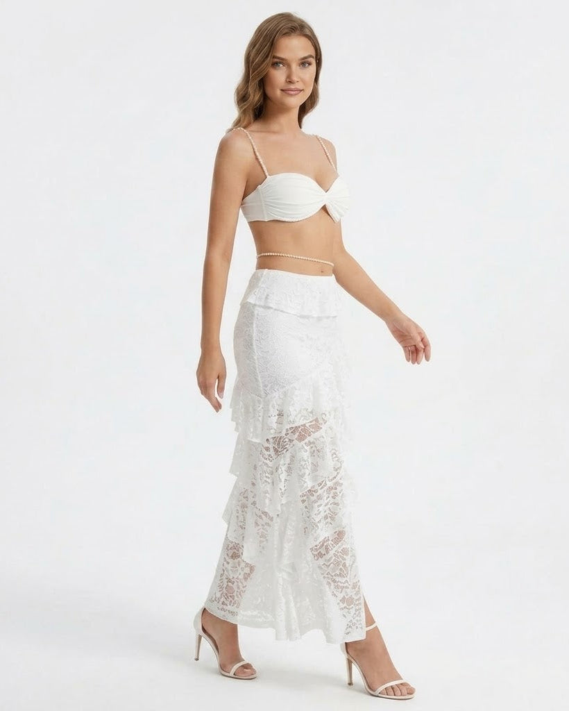 A woman modeling a white two-piece outfit consisting of a ruched bandeau crop top and a high-waisted, tiered lace maxi skirt, paired with clear high-heeled sandals against a plain white studio background.