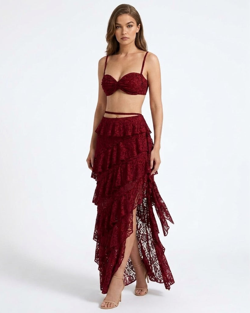 A model wearing a two-piece deep burgundy lace set consisting of a strappy bralette top and a high-low ruffled skirt with a side slit, shot against a plain white studio background.