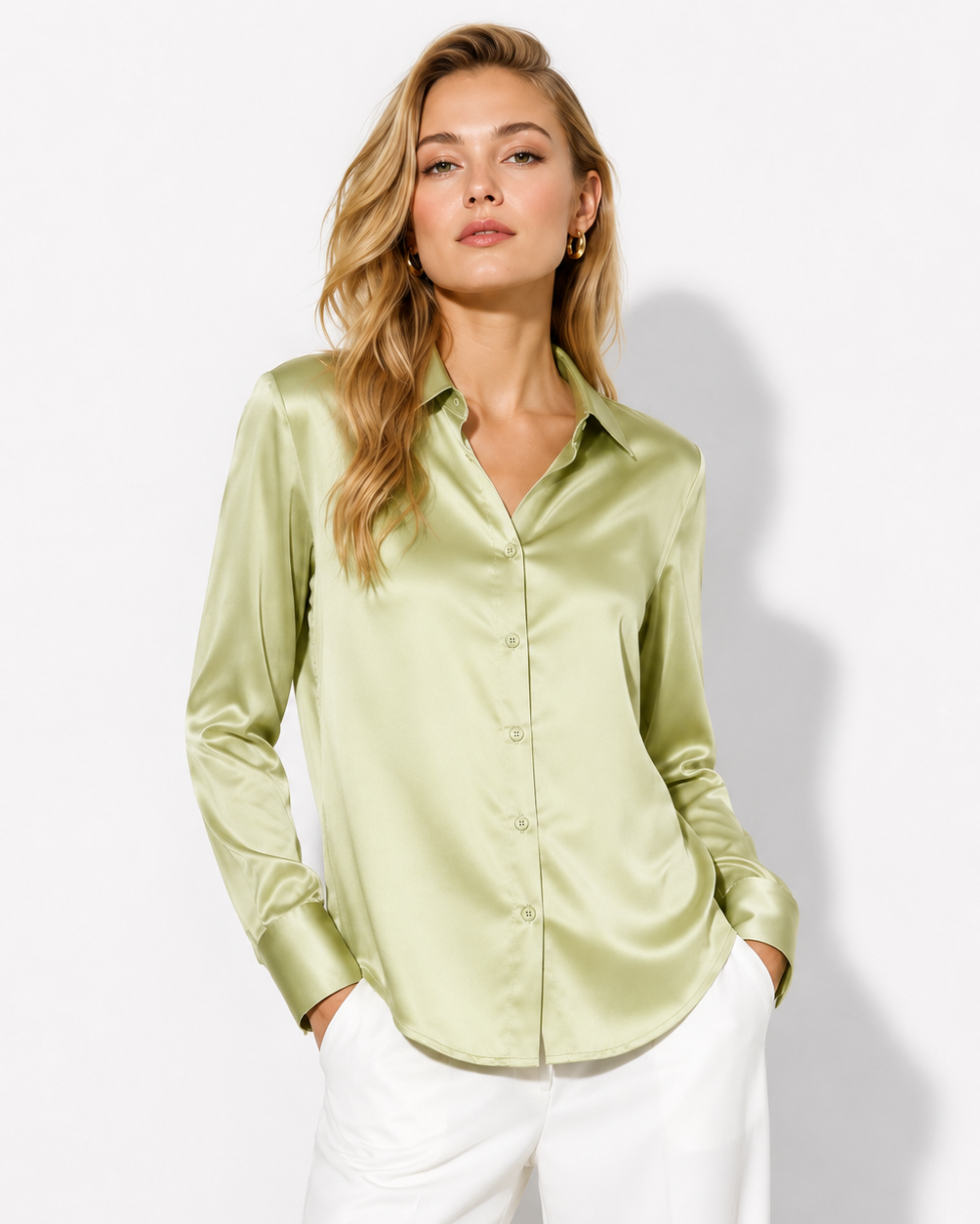 A woman modeling a light olive green satin button-down shirt with white pants, standing against a plain white studio background.