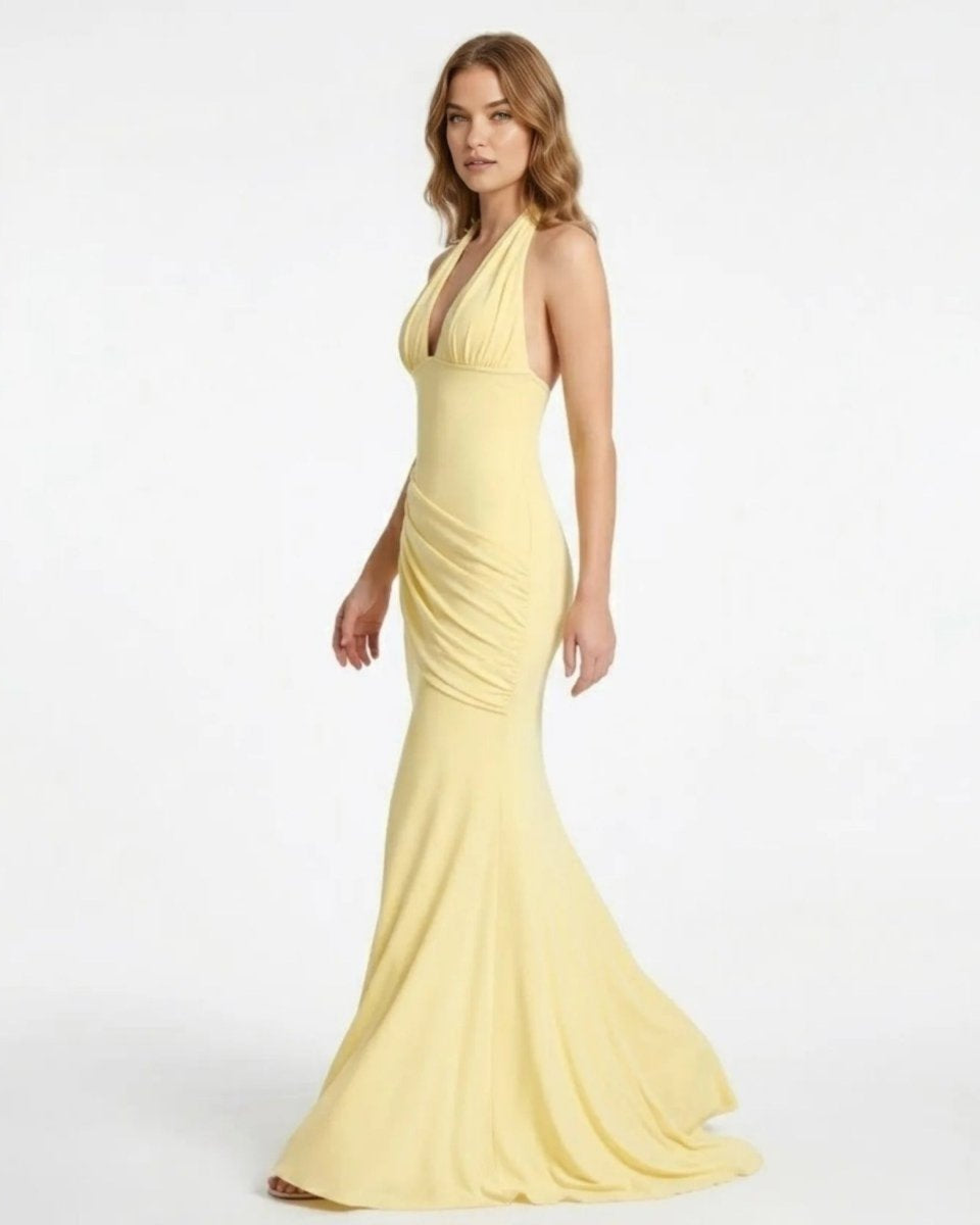 A woman modeling a pale yellow halter-neck mermaid gown with ruched detailing at the waist, photographed against a plain white studio background.