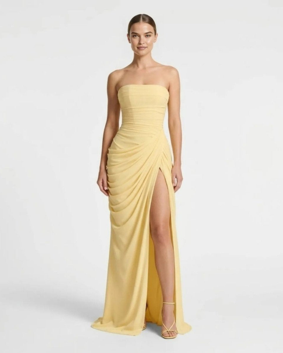 A woman modeling a strapless yellow maxi gown with a ruched bodice, draped skirt, and high front slit, photographed against a clean light gray studio background.
