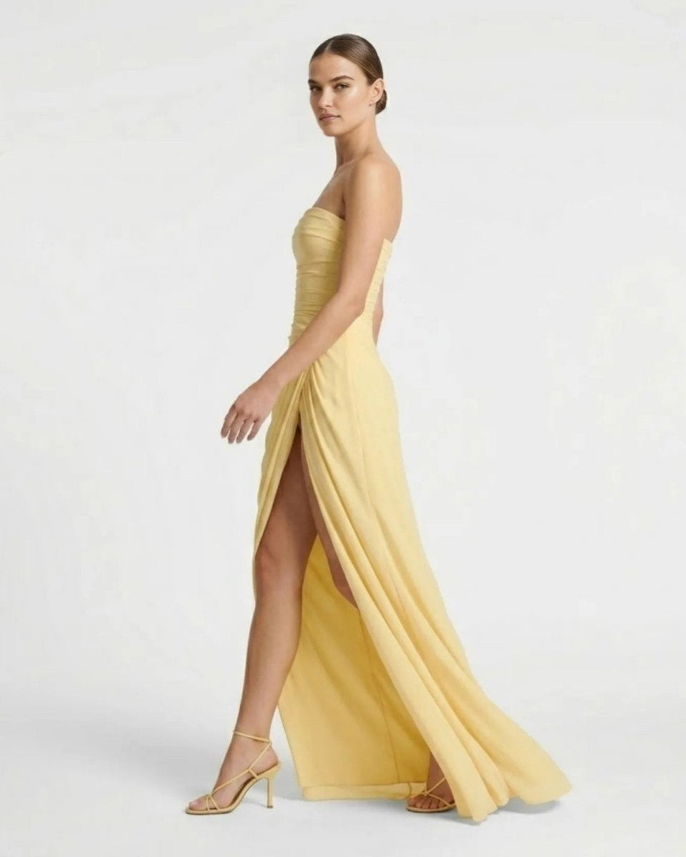 A woman modeling a strapless yellow formal gown with a high thigh slit and flowing skirt, paired with nude high-heel sandals, against a plain light gray studio background.