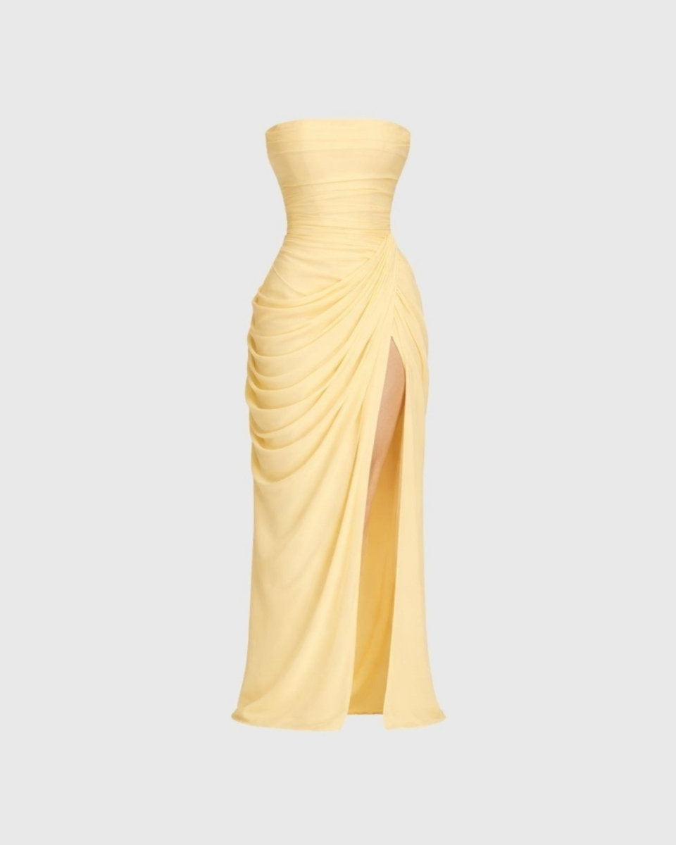 A pale yellow strapless maxi dress with a ruched bodice, draped skirt details, and a high front slit, shown against a light gray background.